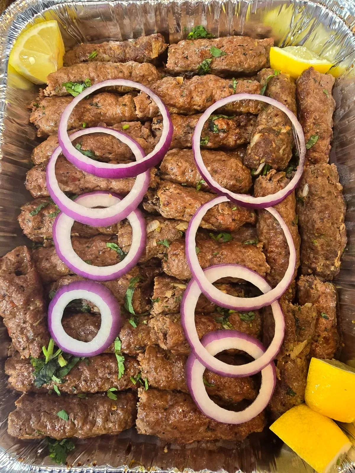 Catering tray of beef seekh kabab garnished with red onion, cilantro and lemon