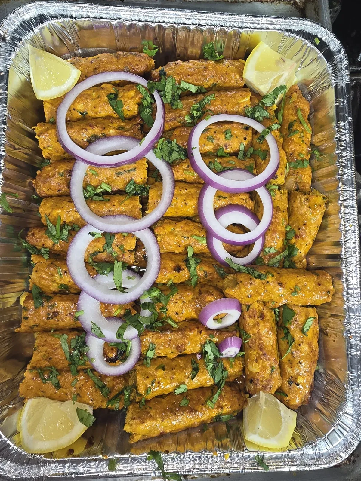 Catering tray of chicken seekh kabab garnished with red onion, cilantro and lemon