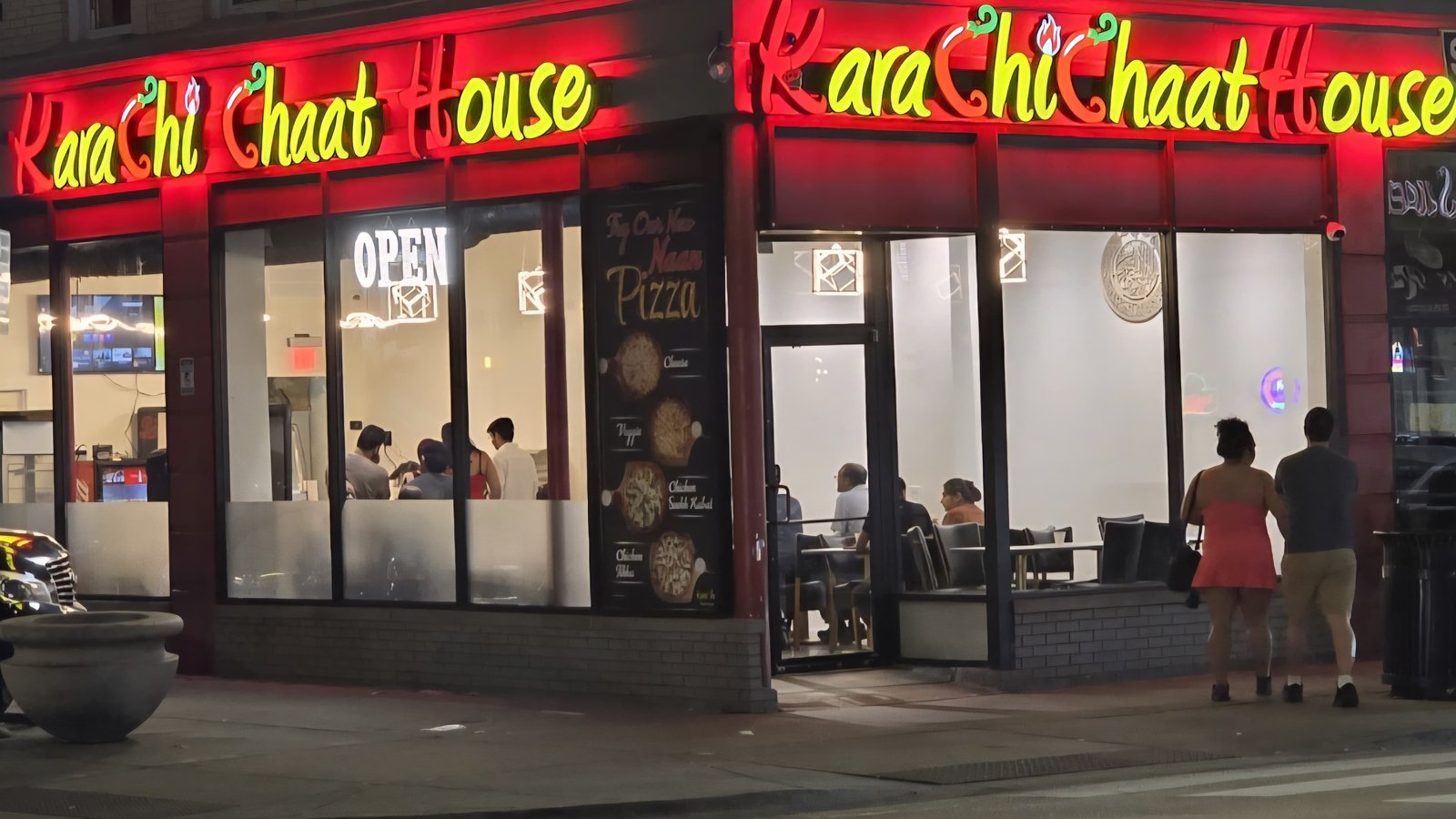 Karachi Chaat House storefront at night on Devon Avenue, Chicago — neon sign glowing red and yellow with diners visible through the window